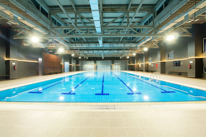 Indoor swimming pool with clear water and bright lighting, surrounded by a modern gym facility.