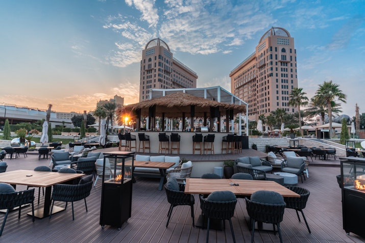 Outdoor lounge area with seating and a bar, framed by two tall buildings during sunset.