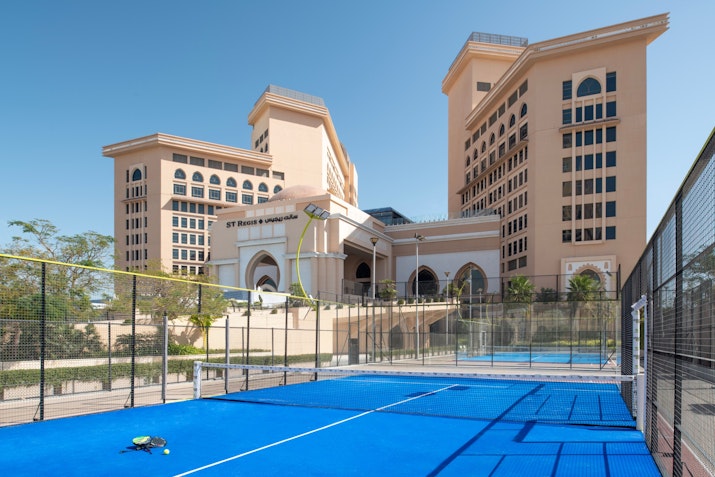 Tennis court with a blue surface, surrounded by fencing and resort buildings in the background