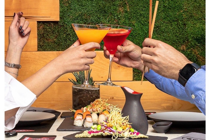Two people toasting with cocktails over a sushi meal.