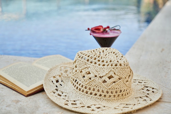 A straw hat, an open book, and a cocktail placed near a swimming pool.