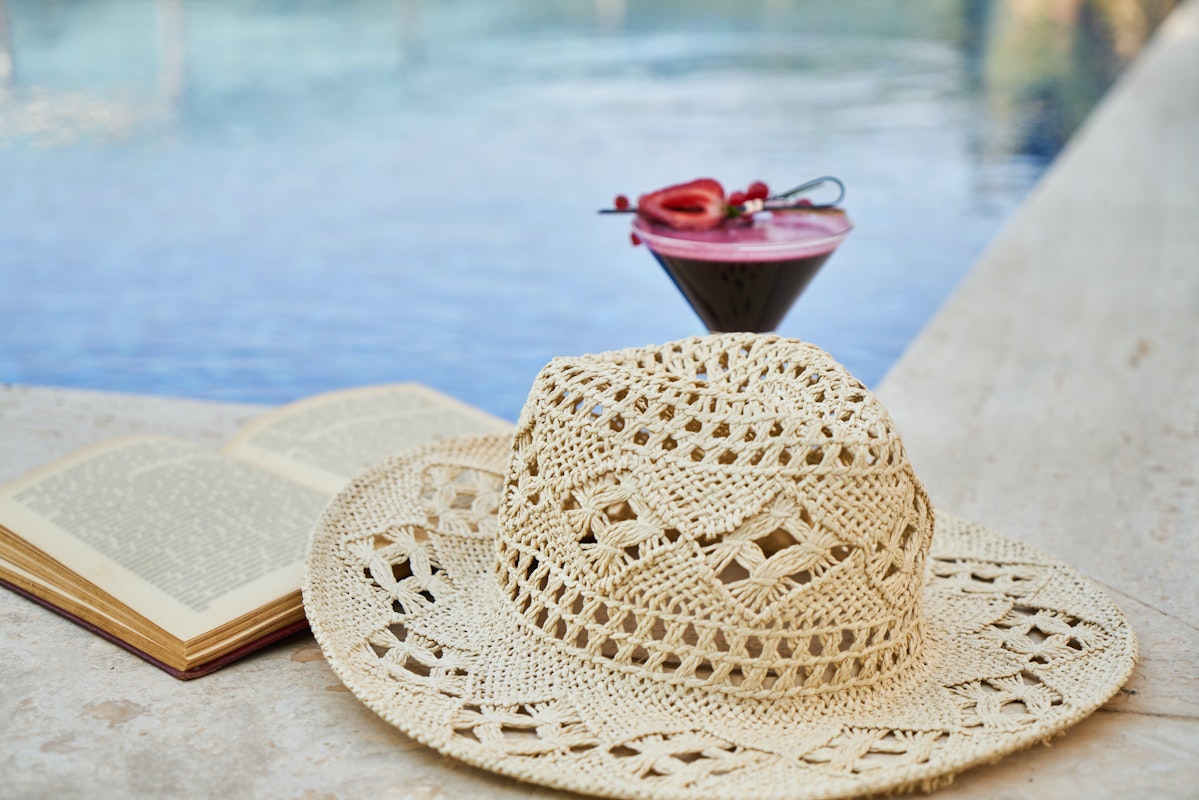 A straw hat, an open book, and a cocktail placed near a swimming pool.