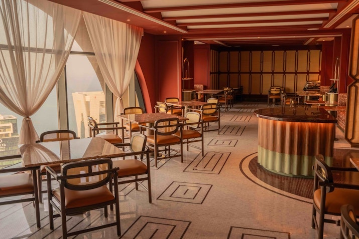 Elegant restaurant interior with round tables, plush seating, and large windows offering a city view.