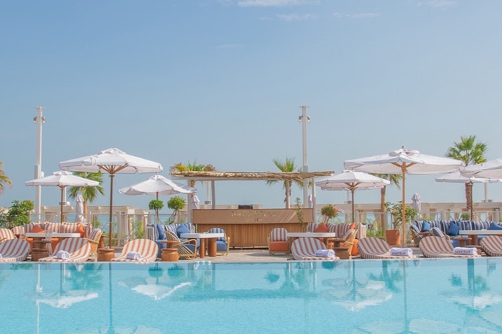 Outdoor pool with lounge chairs, umbrellas, and a bar area, surrounded by clear blue skies