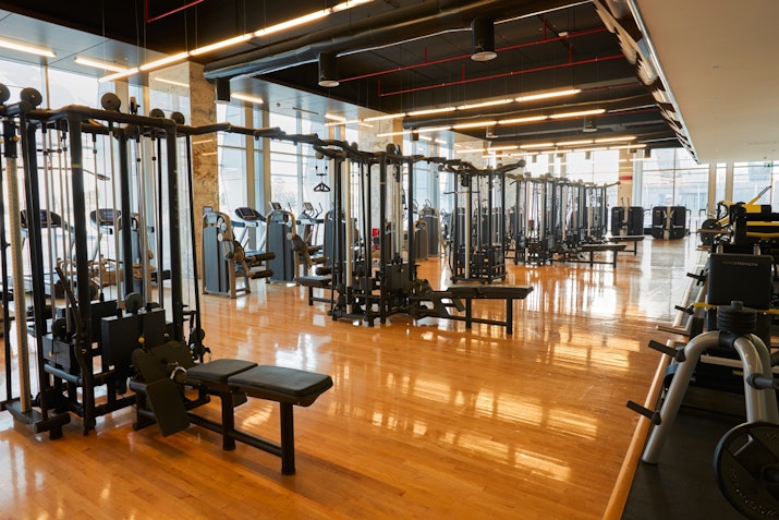 Spacious gym with a variety of exercise equipment and wooden flooring, featuring natural light.