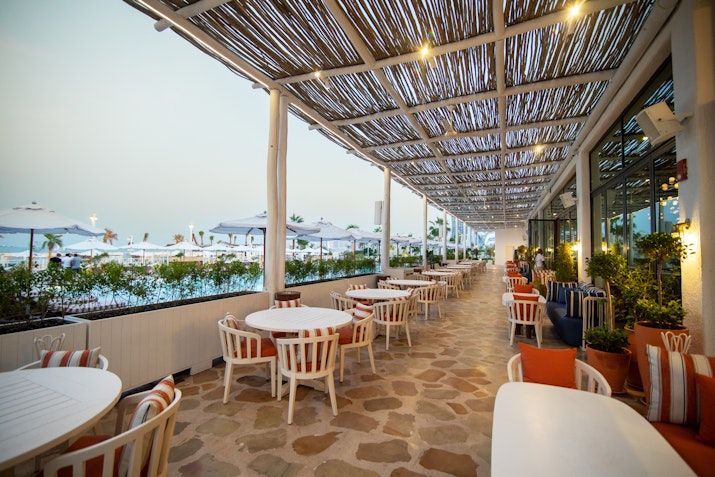 Outdoor dining area with striped chairs and tables, shaded by a woven canopy, overlooking a pool area with umbrellas and plants.
