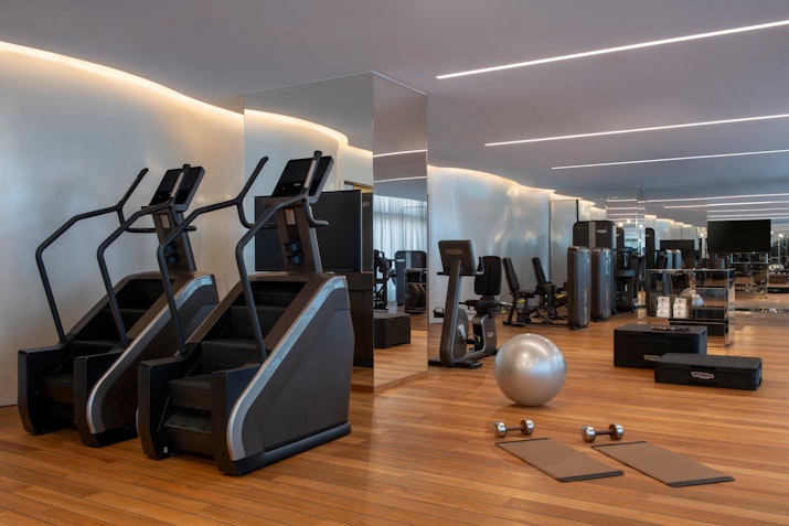 Modern gym with ellipticals, exercise machines, dumbbells, and mats on a wooden floor.