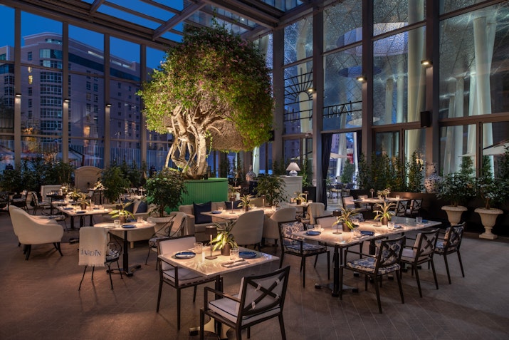 Elegant restaurant with dining tables and a large tree in the center, surrounded by plants and ambient lighting