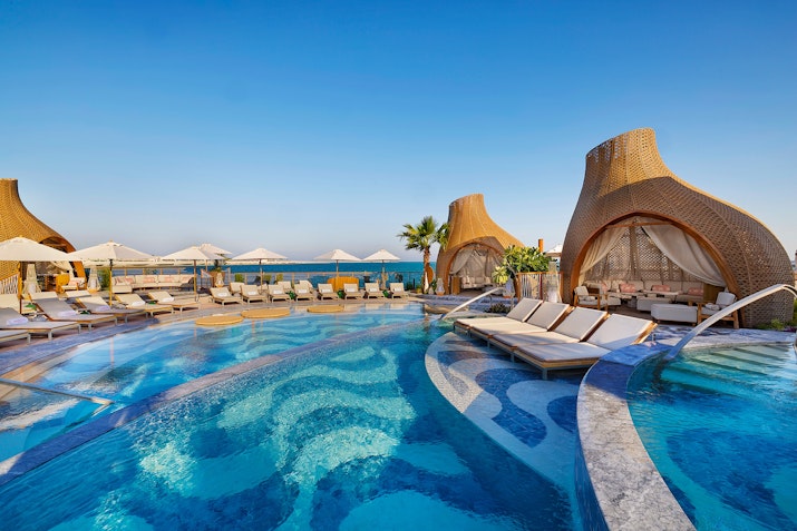 A luxurious pool area with cabanas, lounge chairs, and a clear blue sky overlooking the sea.