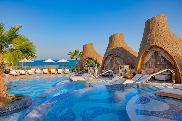An elegant pool area featuring three cabanas with water features, surrounded by lounge chairs, with a scenic view of the ocean under a clear blue sky.