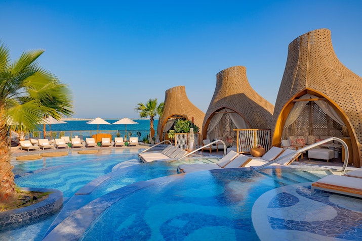 An elegant pool area featuring three cabanas with water features, surrounded by lounge chairs, with a scenic view of the ocean under a clear blue sky.