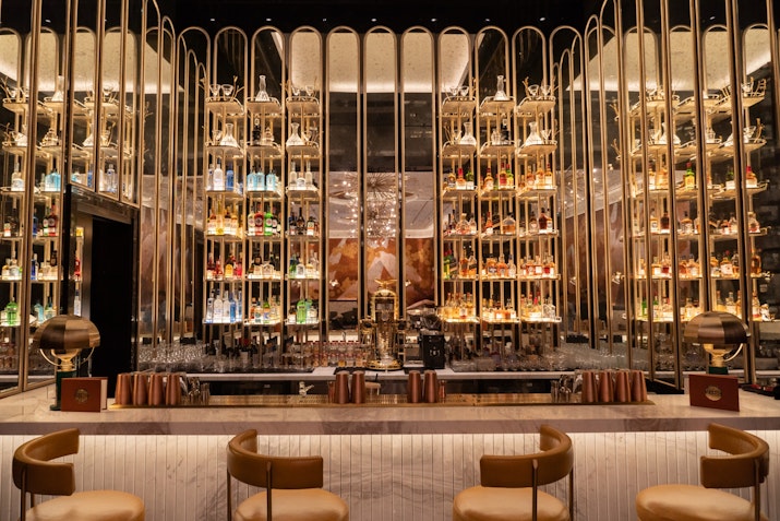 Luxurious bar with shelves of liquor bottles, illuminated by soft lighting, and elegant bar stools.