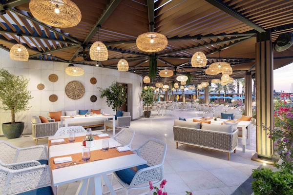 Outdoor dining area with tables set up under hanging lights, surrounded by plants and modern furniture.