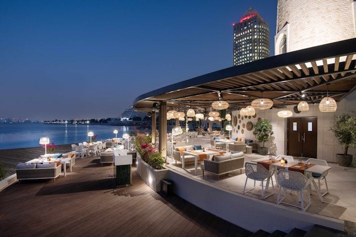 Outdoor restaurant with cozy seating and ambient lighting overlooking the water at night, with a city skyline in the background.