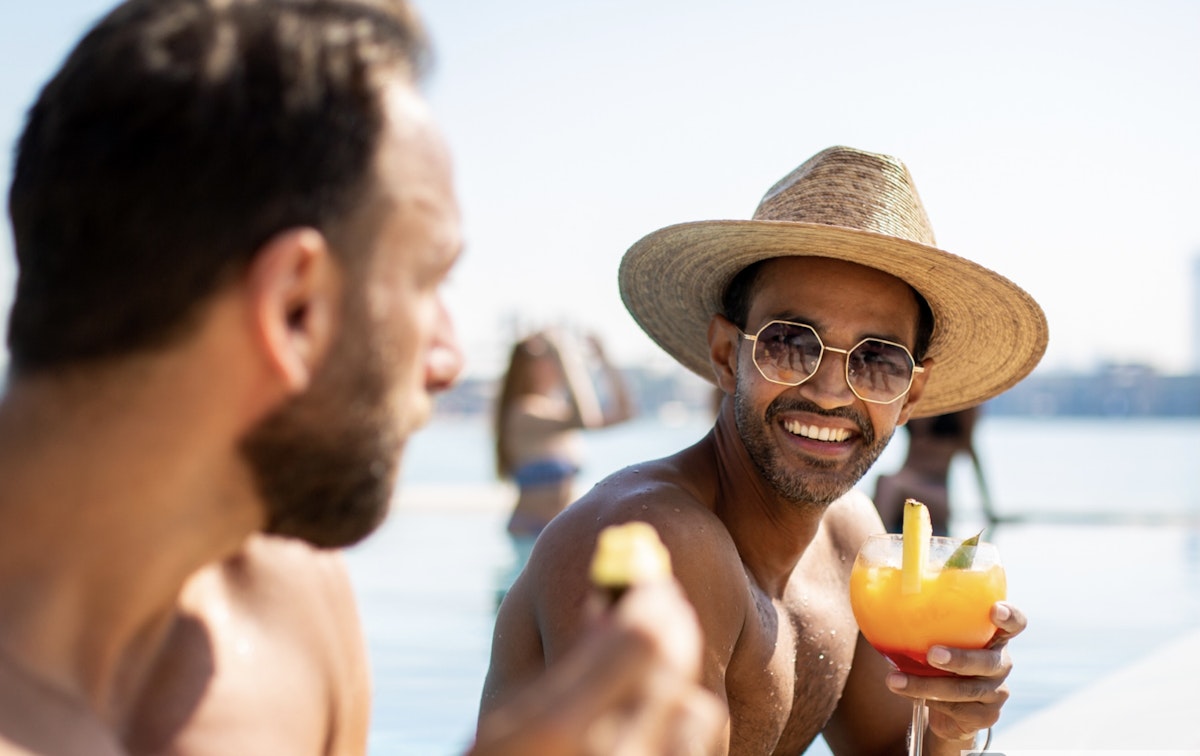 A man smiling while holding a tropical drink, wearing sunglasses and a straw hat, with a person in the background.