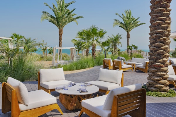 Praia at Waldorf Astoria Lusail Relaxing outdoor seating area with wooden and white chairs surrounded by palm trees, with a view of the beach.