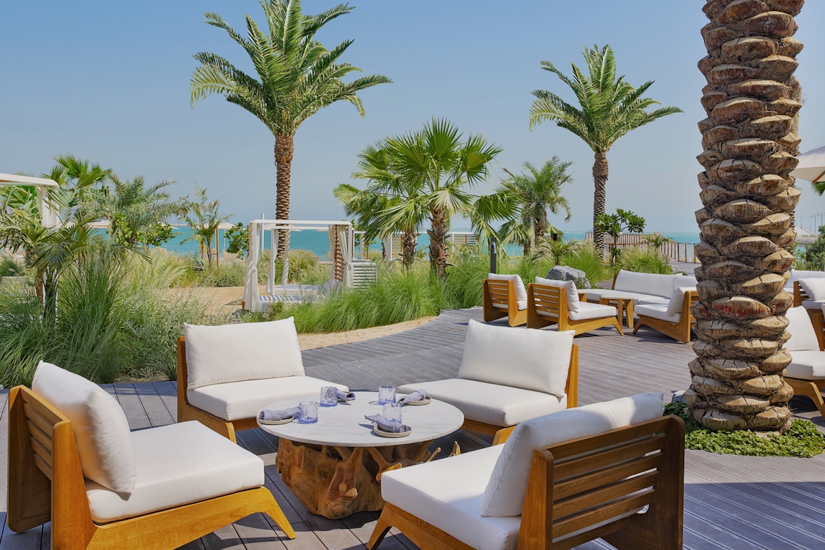 Praia at Waldorf Astoria Lusail Relaxing outdoor seating area with wooden and white chairs surrounded by palm trees, with a view of the beach.
