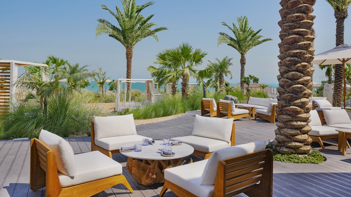 Relaxing outdoor seating area with wooden and white chairs surrounded by palm trees, with a view of the beach.
