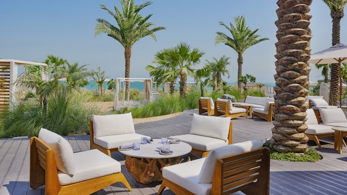 Relaxing outdoor seating area with wooden and white chairs surrounded by palm trees, with a view of the beach.