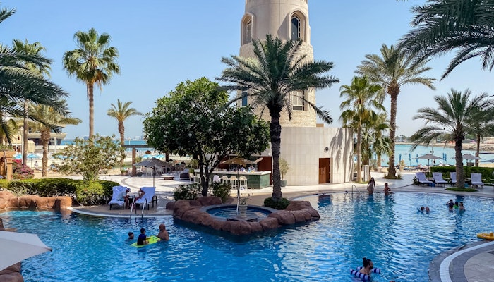 The Four Seasons Hotel Doha Resort pool with people swimming, palm trees, and a tower in the background.