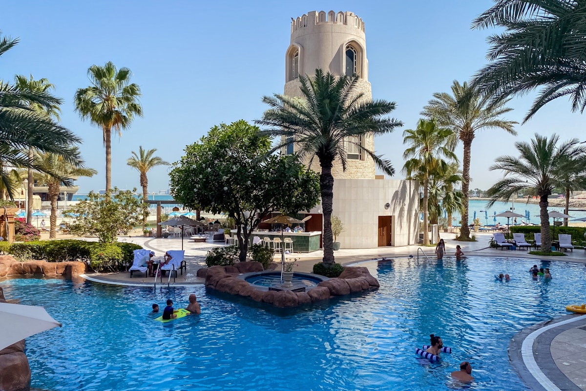 The Four Seasons Hotel Doha Resort pool with people swimming, palm trees, and a tower in the background.