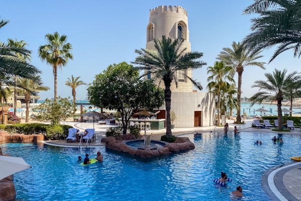Resort pool with people swimming, palm trees, and a tower in the background.
