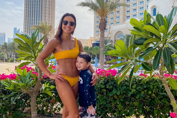 Woman in a yellow bikini with a child standing together in front of plants and a hotel