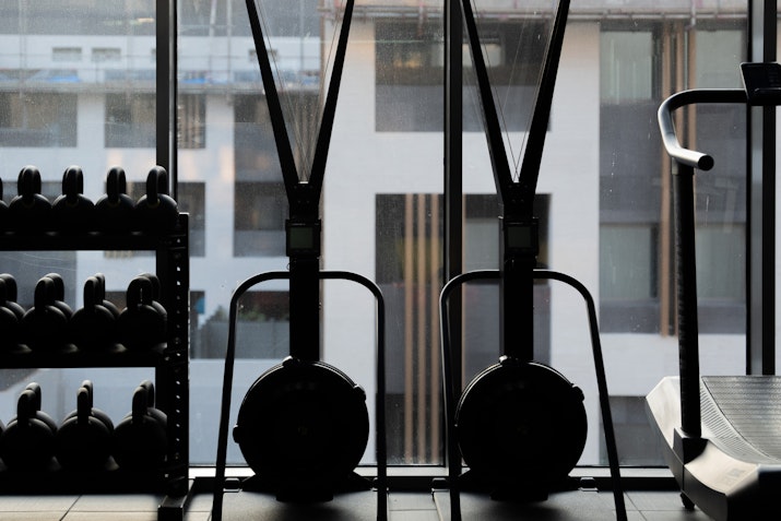 Two gym rowing machines with kettlebells stacked beside them, visible through large windows