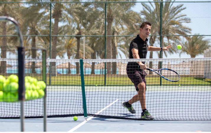 Tennis player hitting a ball on the court with a tennis racket, with palm trees in the background.
