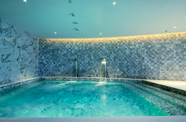 Indoor spa pool with water fountains, illuminated by soft lighting and surrounded by marble walls.