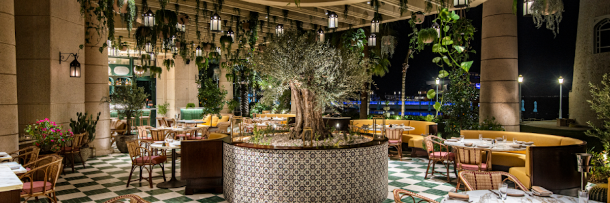 Take your tastebuds on an adventure! Outdoor dining area with hanging plants and a fountain, offering a cozy atmosphere with a checkered floor.