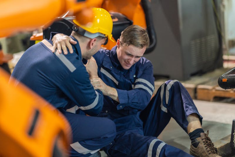A man has been injured by one of the machines he works with. He has injured his leg and another employee is trying to lift him off the ground. Both men are wearing blue work suits and there is a lare orange machine behind them.