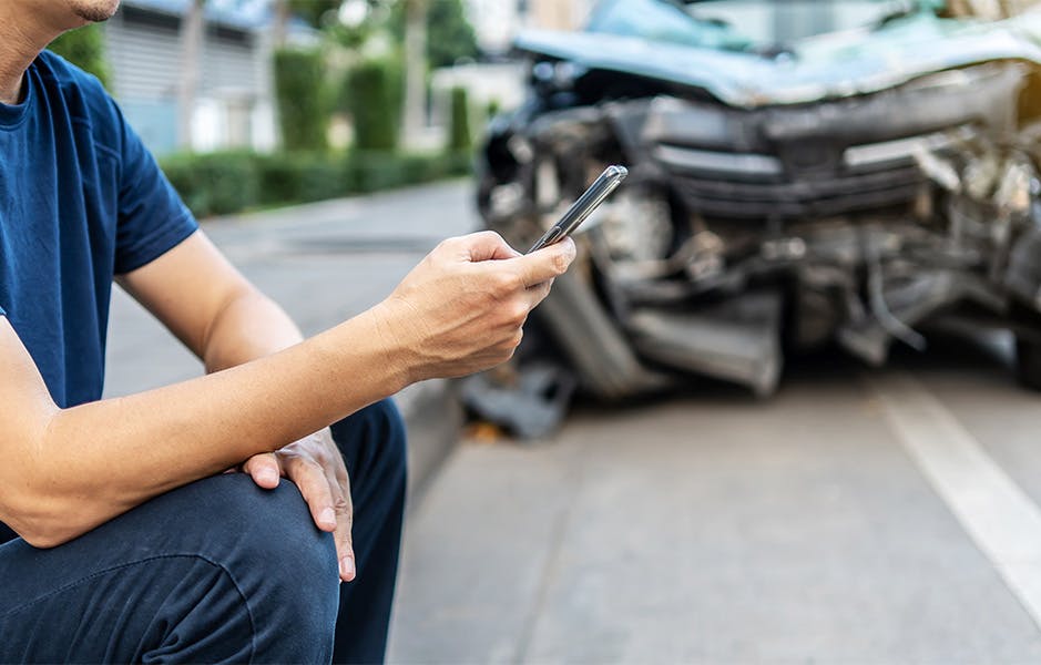man sitting on the curb with phone in hand with car crash next to him