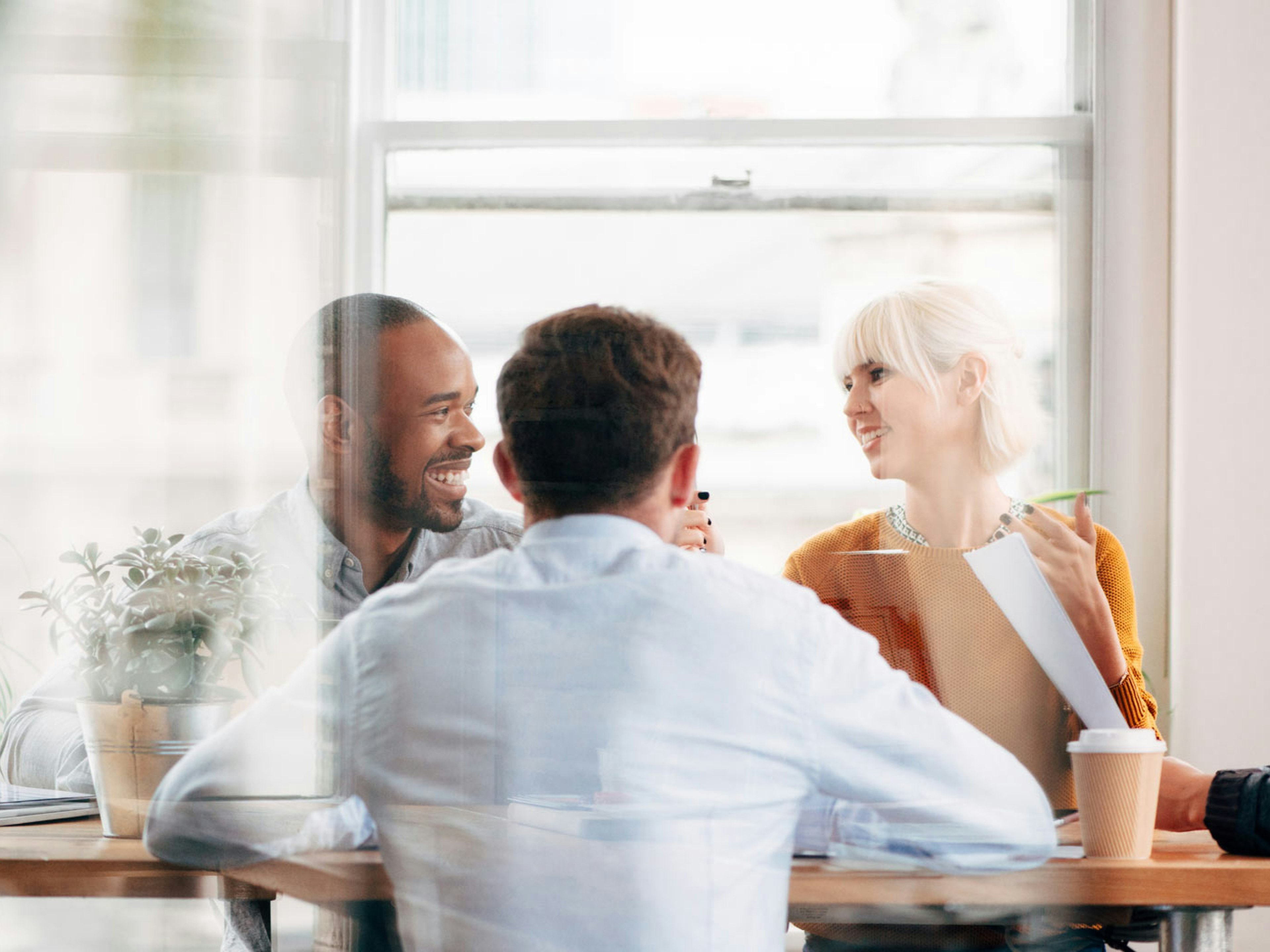 Vier Personen im Gespräch am Tisch, lockere Atmosphäre mit Pflanzen und Kaffeebecher im Büro.