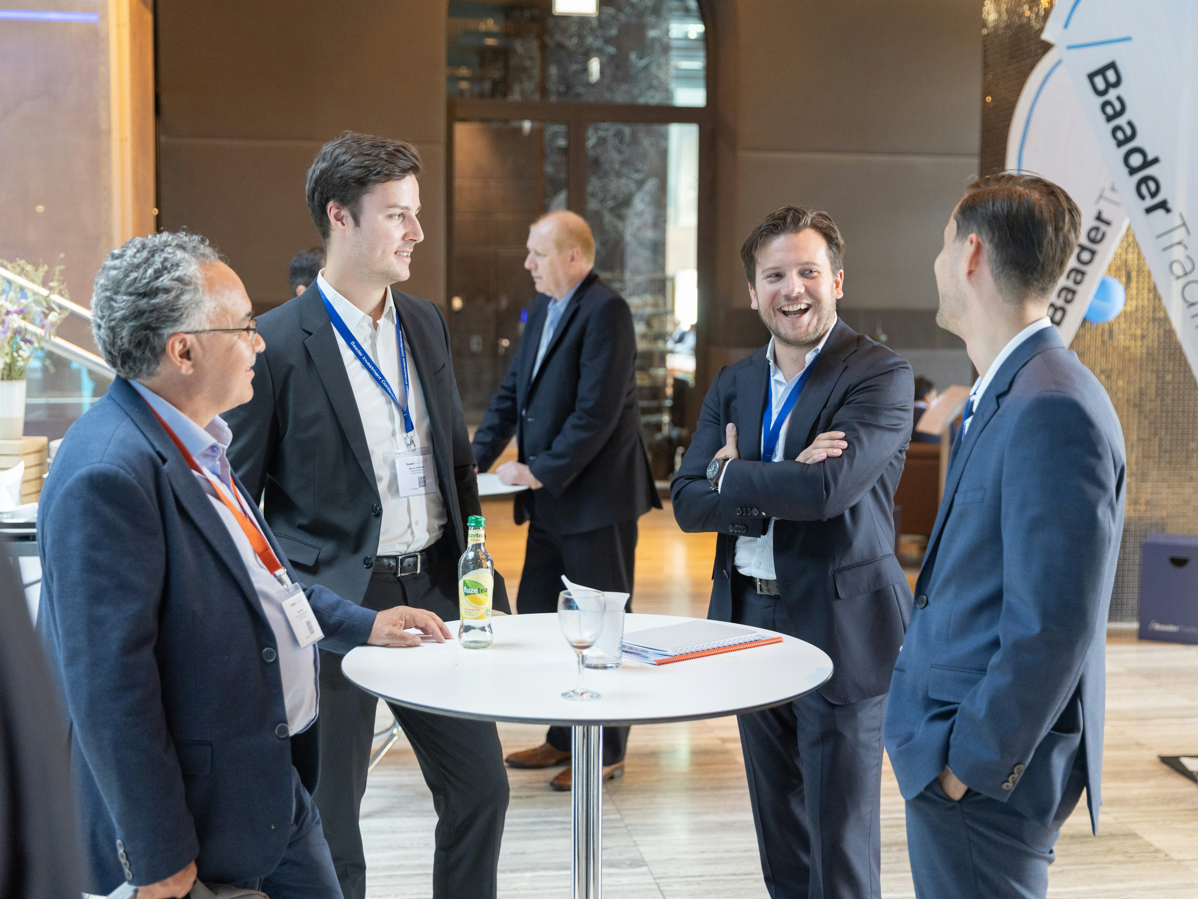 Vier Männer im Anzug stehen lachend an einem Stehtisch bei einer Baader-Bank-Veranstaltung.