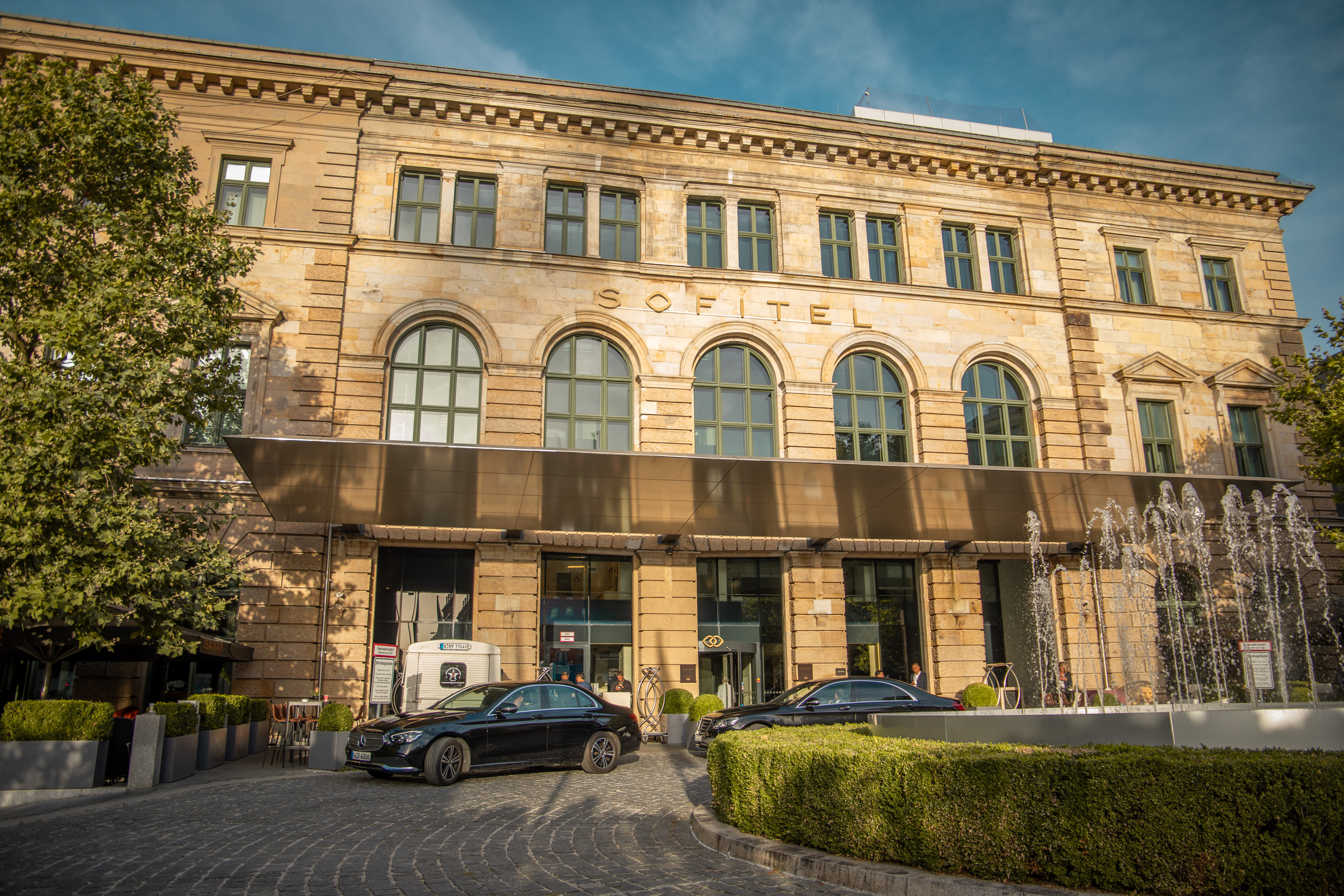 Außenansicht des Sofitel-Hotels, ein mehrstöckiges Sandsteingebäude mit großen Fenstern. Vor dem Eingang stehen mehrere schwarze Autos, daneben ist ein Springbrunnen mit Wasserfontänen zu sehen.