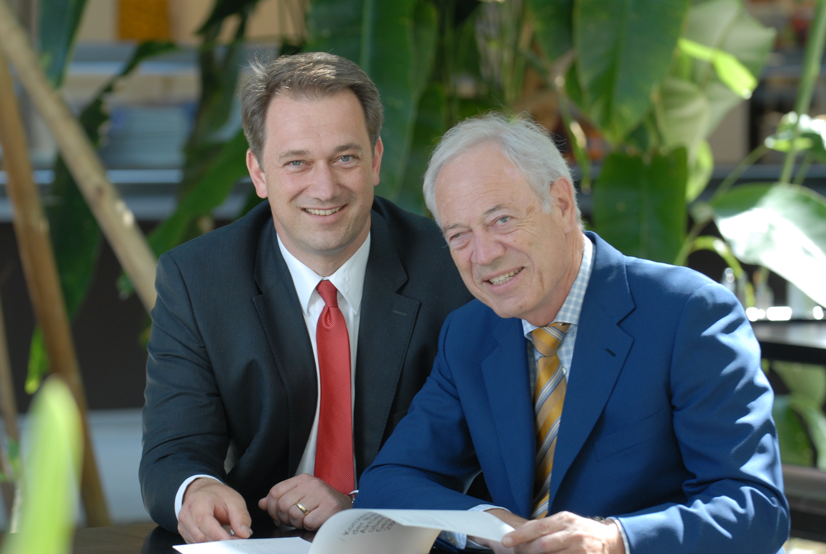 Zwei Männer im Anzug sitzen nebeneinander und blicken läch.elnd in die Kamera