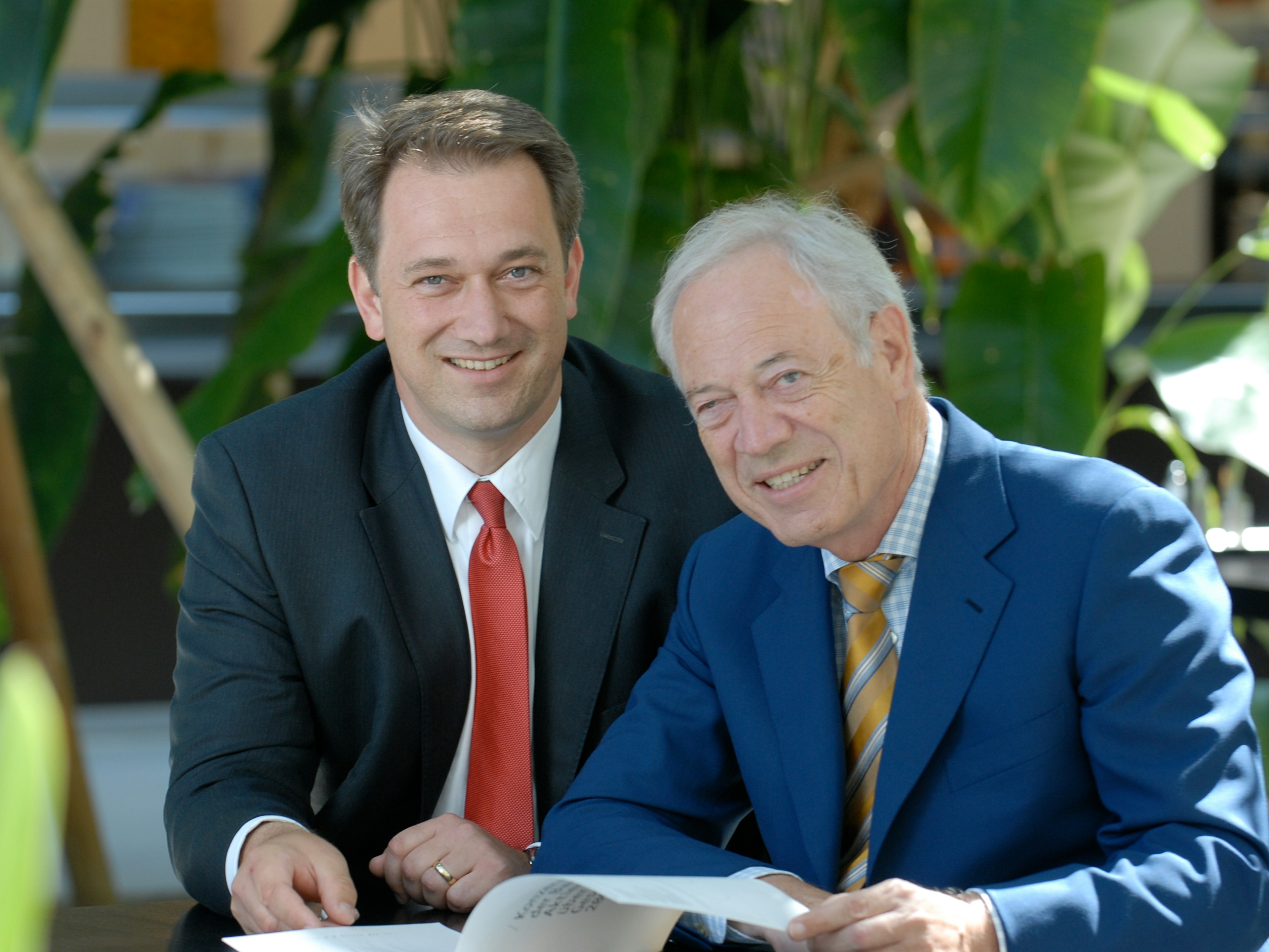 Zwei Männer im Anzug sitzen nebeneinander und blicken läch.elnd in die Kamera