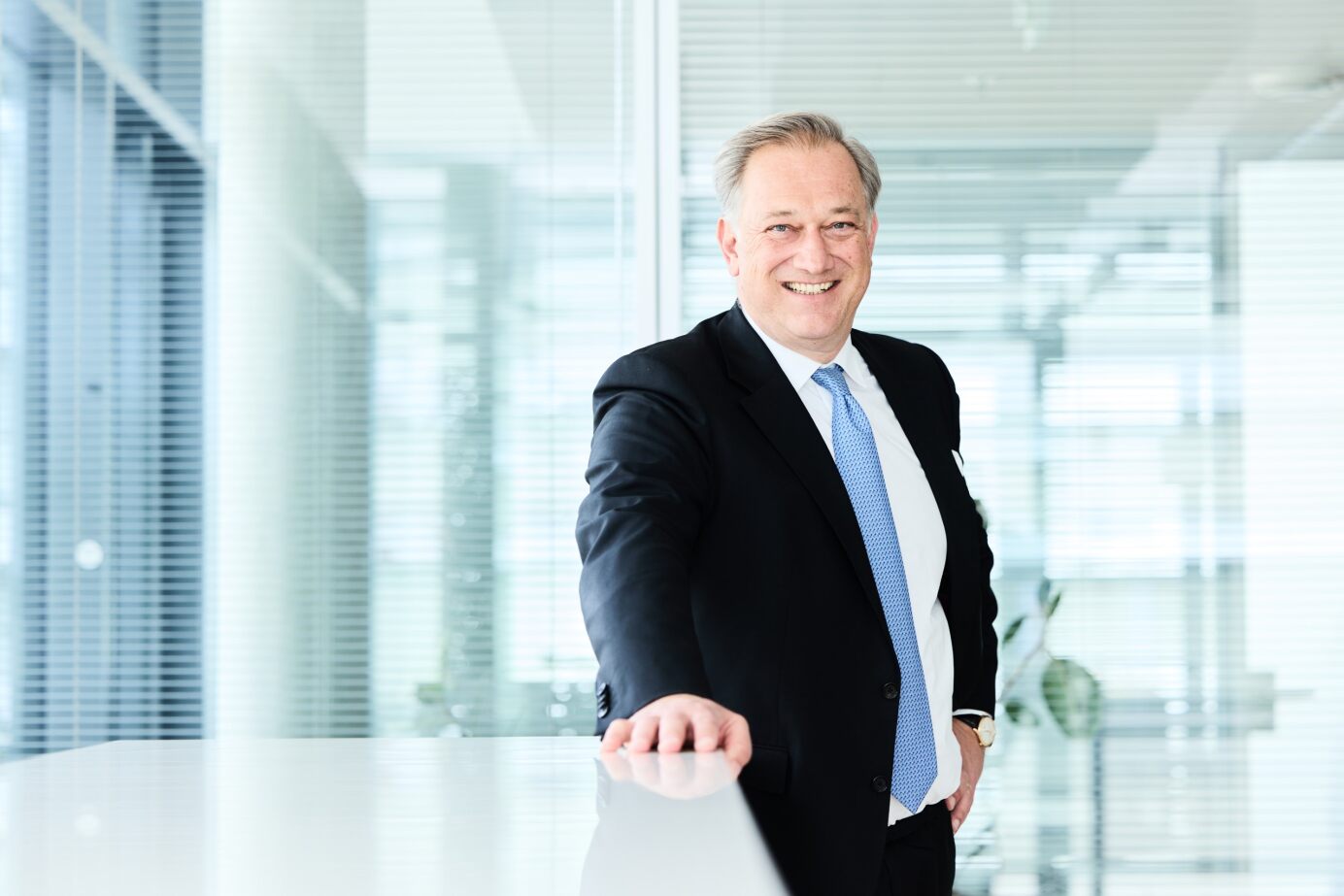 Mann im Anzug mit hellblauer Krawatte steht lächelnd in einem modernen Büro mit Glaswänden.