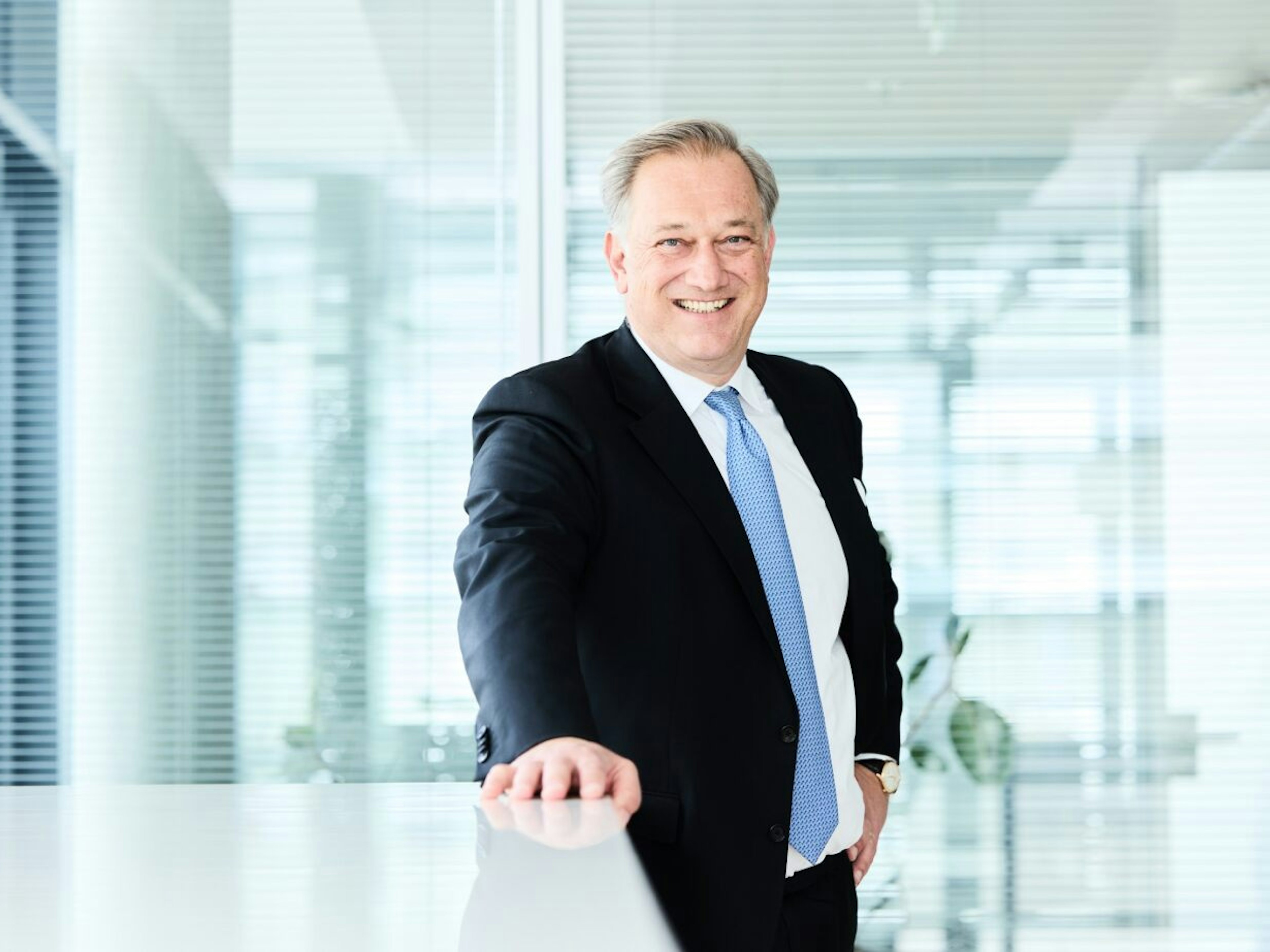 Mann im Anzug mit hellblauer Krawatte steht lächelnd in einem modernen Büro mit Glaswänden.