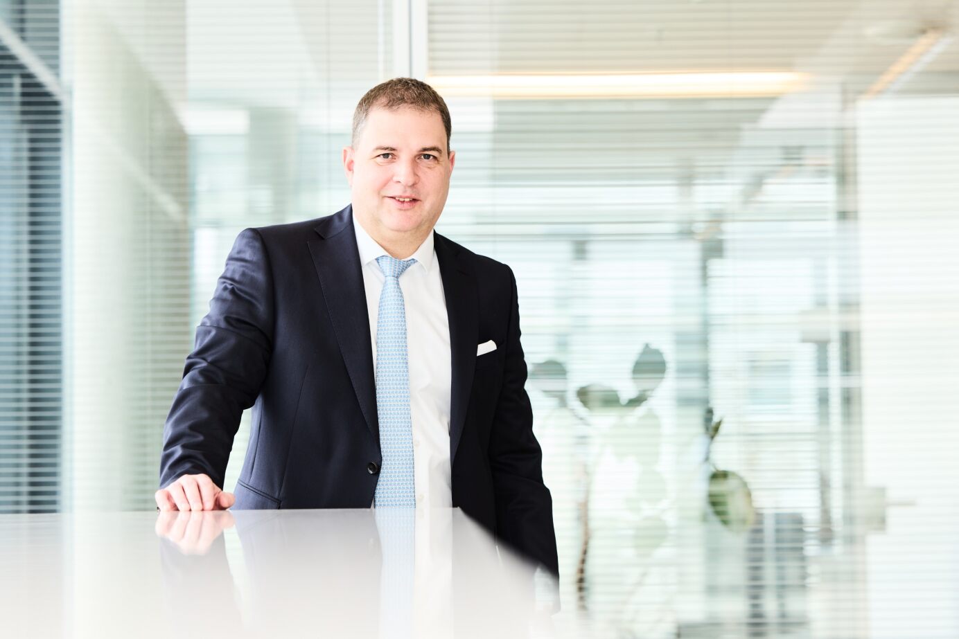 Mann im dunklen Anzug mit hellblauer Krawatte steht lächelnd an einem Tisch in einem modernen Büro mit Glaswänden.