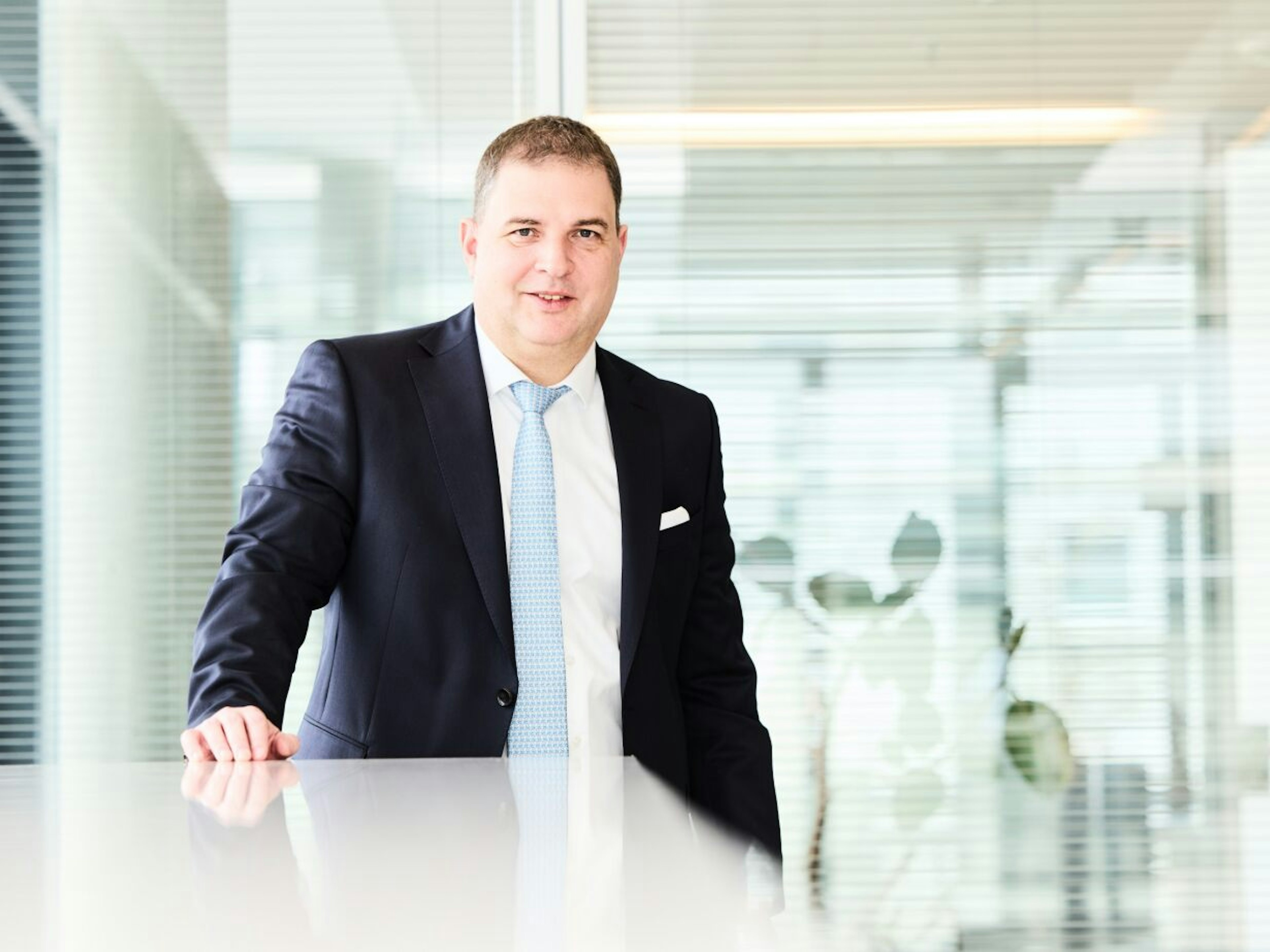 Mann im dunklen Anzug mit hellblauer Krawatte steht lächelnd an einem Tisch in einem modernen Büro mit Glaswänden.