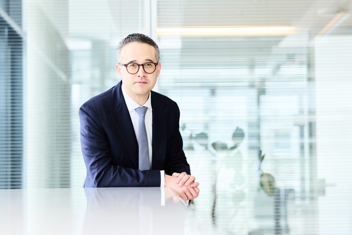 Mann mit Brille und Anzug sitzt an einem Tisch, die Hände gefaltet, Hintergrund mit Glasfront und Pflanzen.