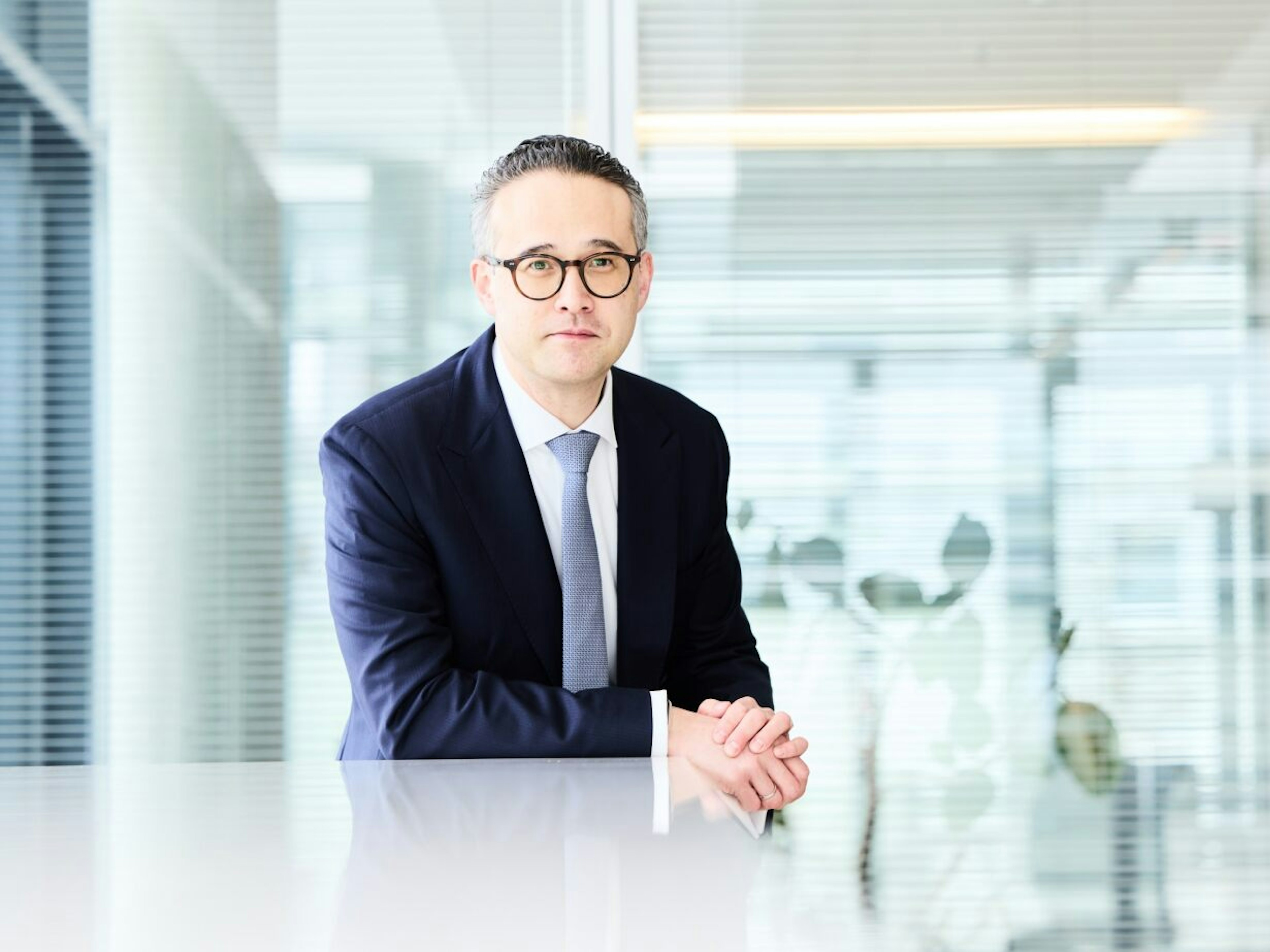 Mann mit Brille und Anzug sitzt an einem Tisch, die Hände gefaltet, Hintergrund mit Glasfront und Pflanzen.