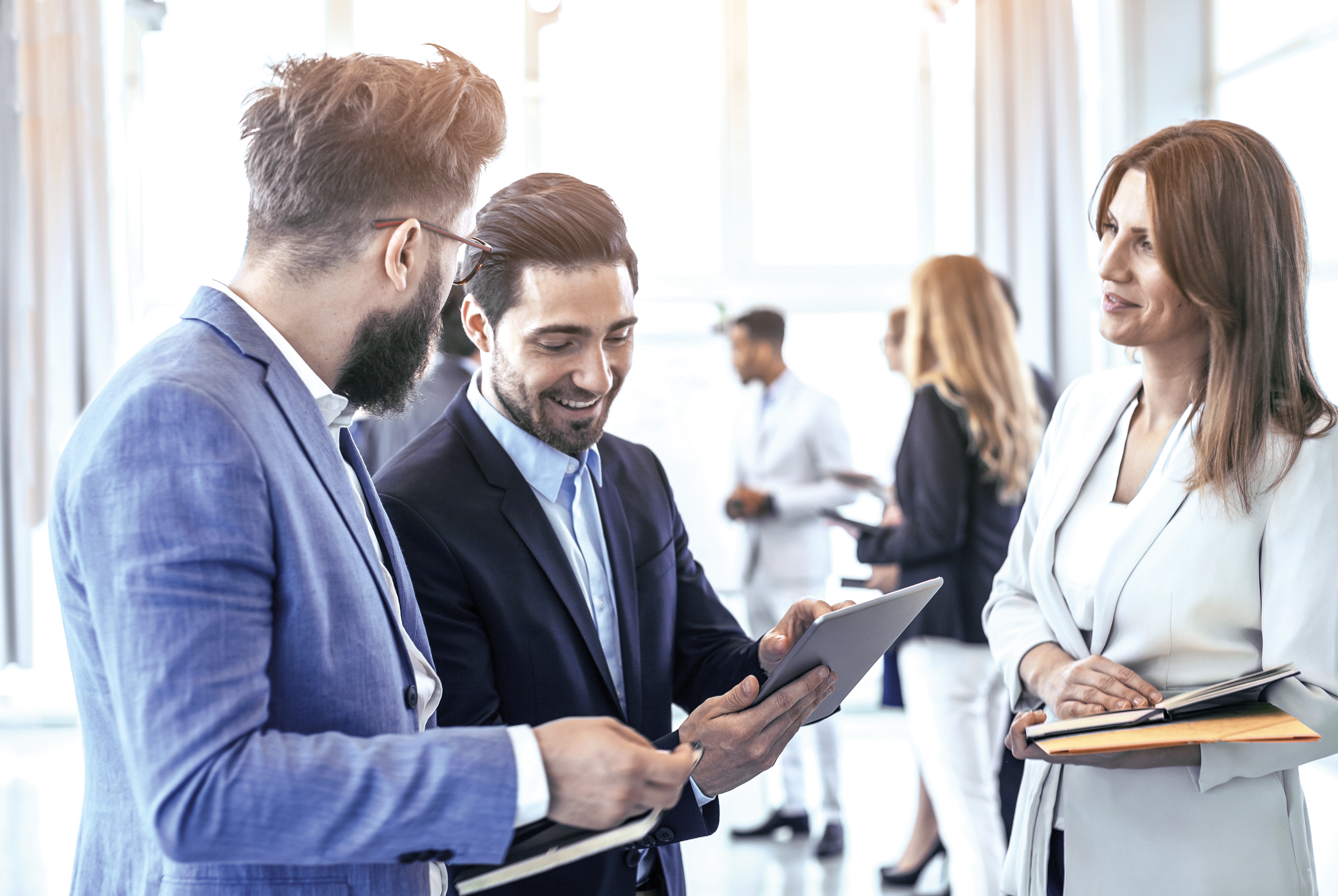 Zwei Männer in Anzügen betrachten gemeinsam ein Tablet und lächeln, daneben steht eine Frau in heller Businesskleidung mit einem Notizbuch in der Hand, im Hintergrund weitere Personen in Business-Umgebung.