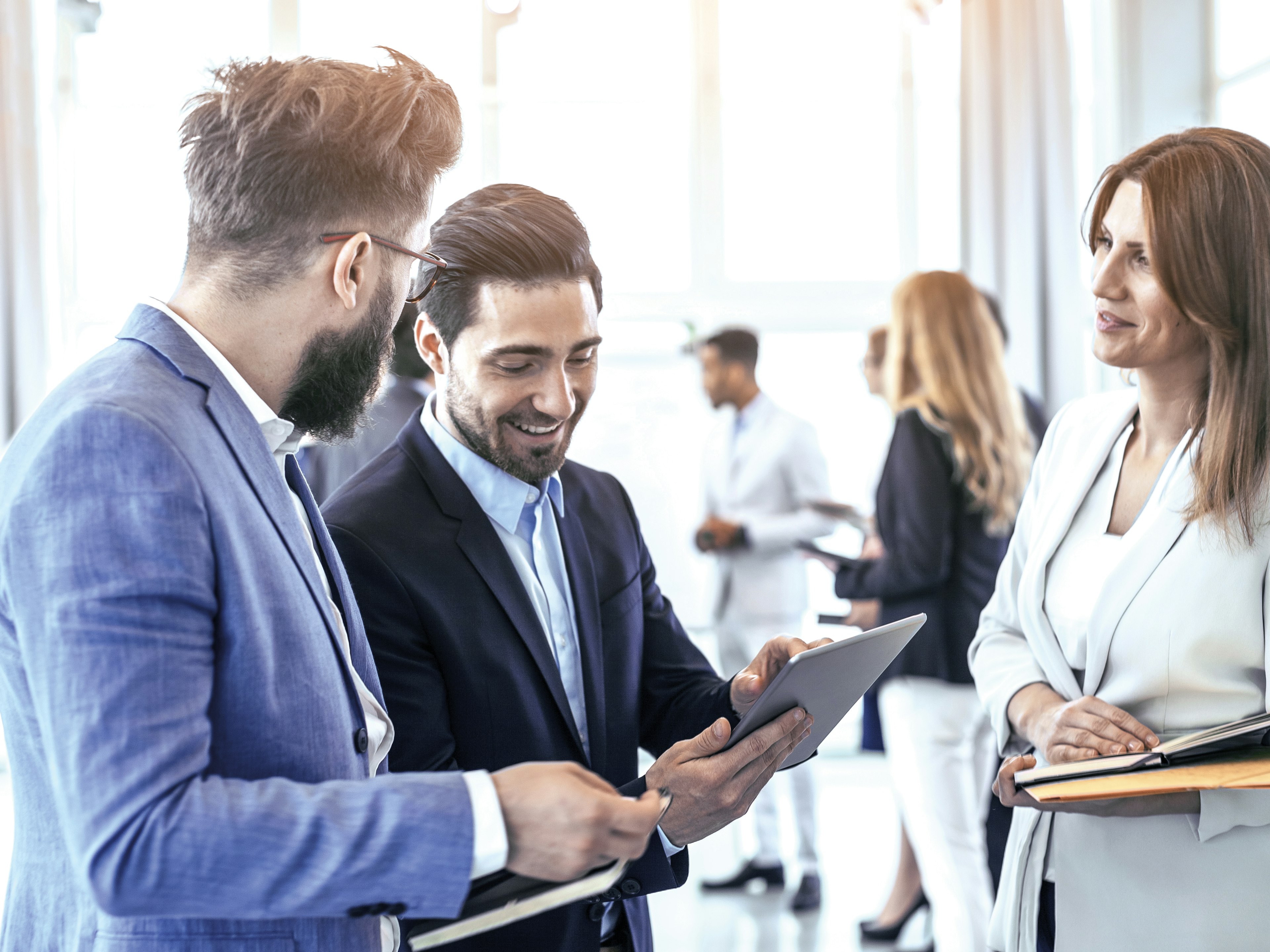 Zwei Männer in Anzügen betrachten gemeinsam ein Tablet und lächeln, daneben steht eine Frau in heller Businesskleidung mit einem Notizbuch in der Hand, im Hintergrund weitere Personen in Business-Umgebung.