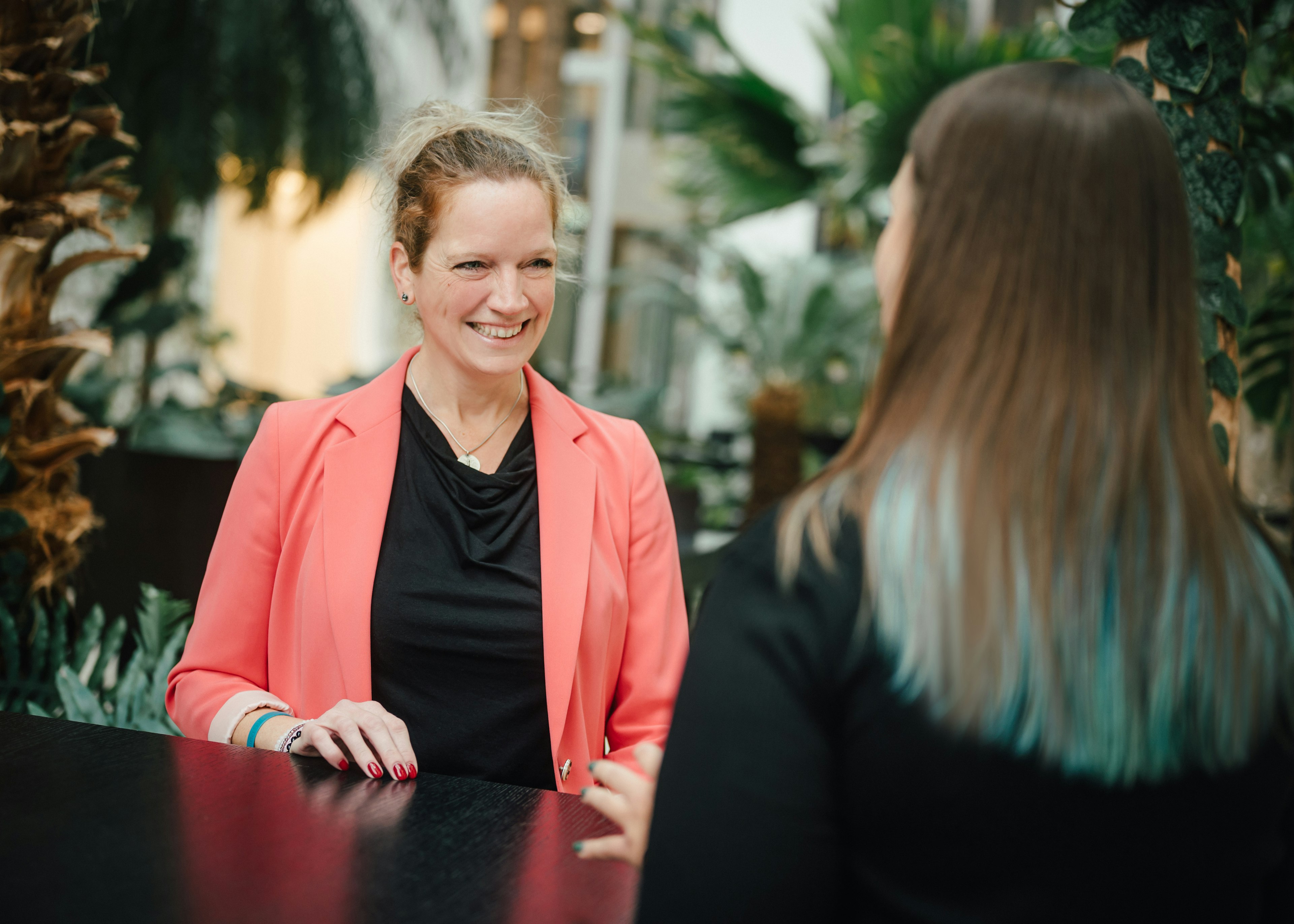 Eine Frau mit hochgesteckten Haaren und pinkfarbenem Blazer sitzt lächelnd an einem Tisch und spricht mit einer Frau, die von hinten zu sehen ist und schulterlanges Haar mit blauen Strähnen trägt, im Hintergrund sind Pflanzen und Glaswände sichtbar.