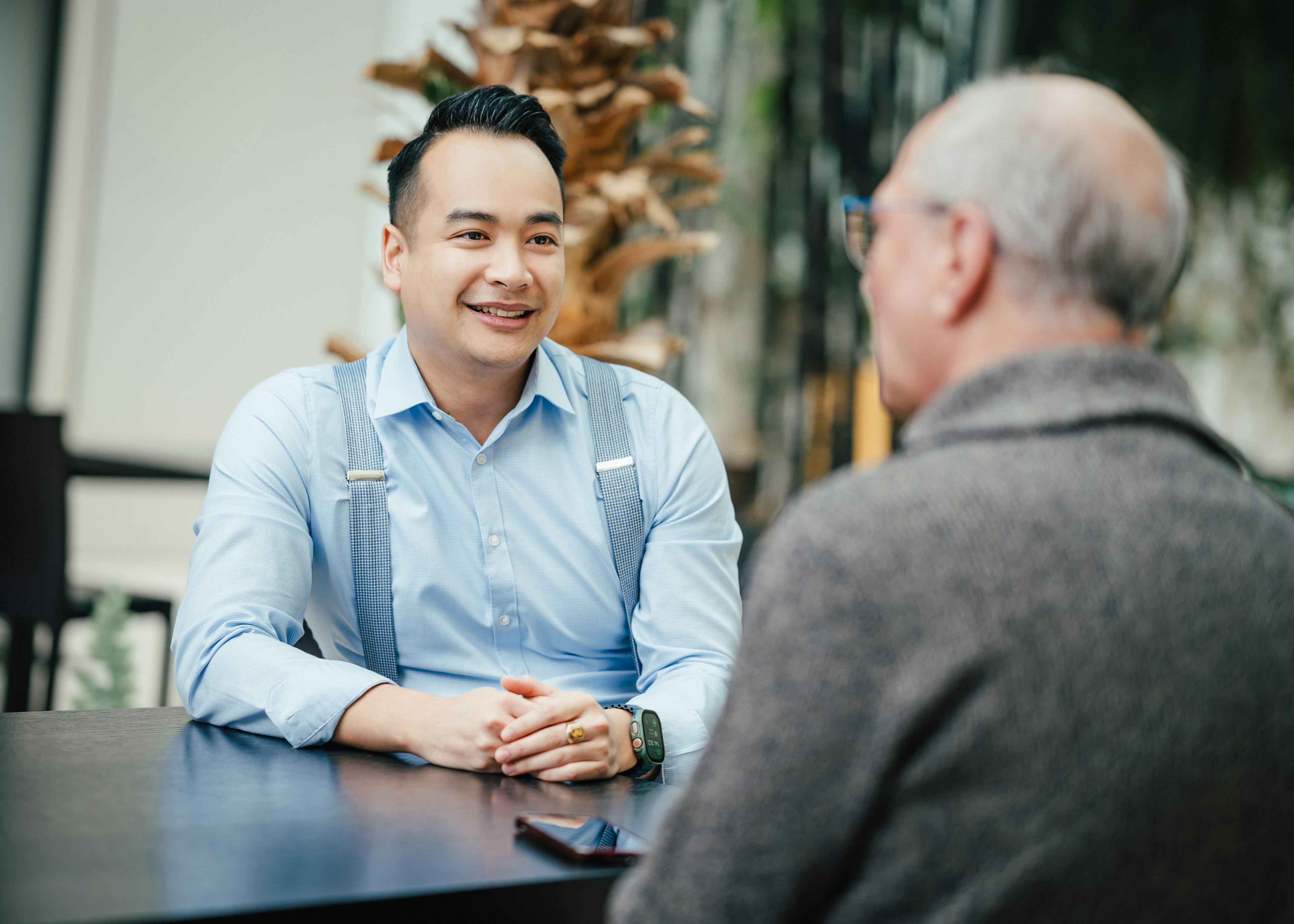 Zwei Männer sitzen an einem Tisch und sprechen miteinander, einer trägt Hemd mit Hosenträgern, der andere grauen Pullover.