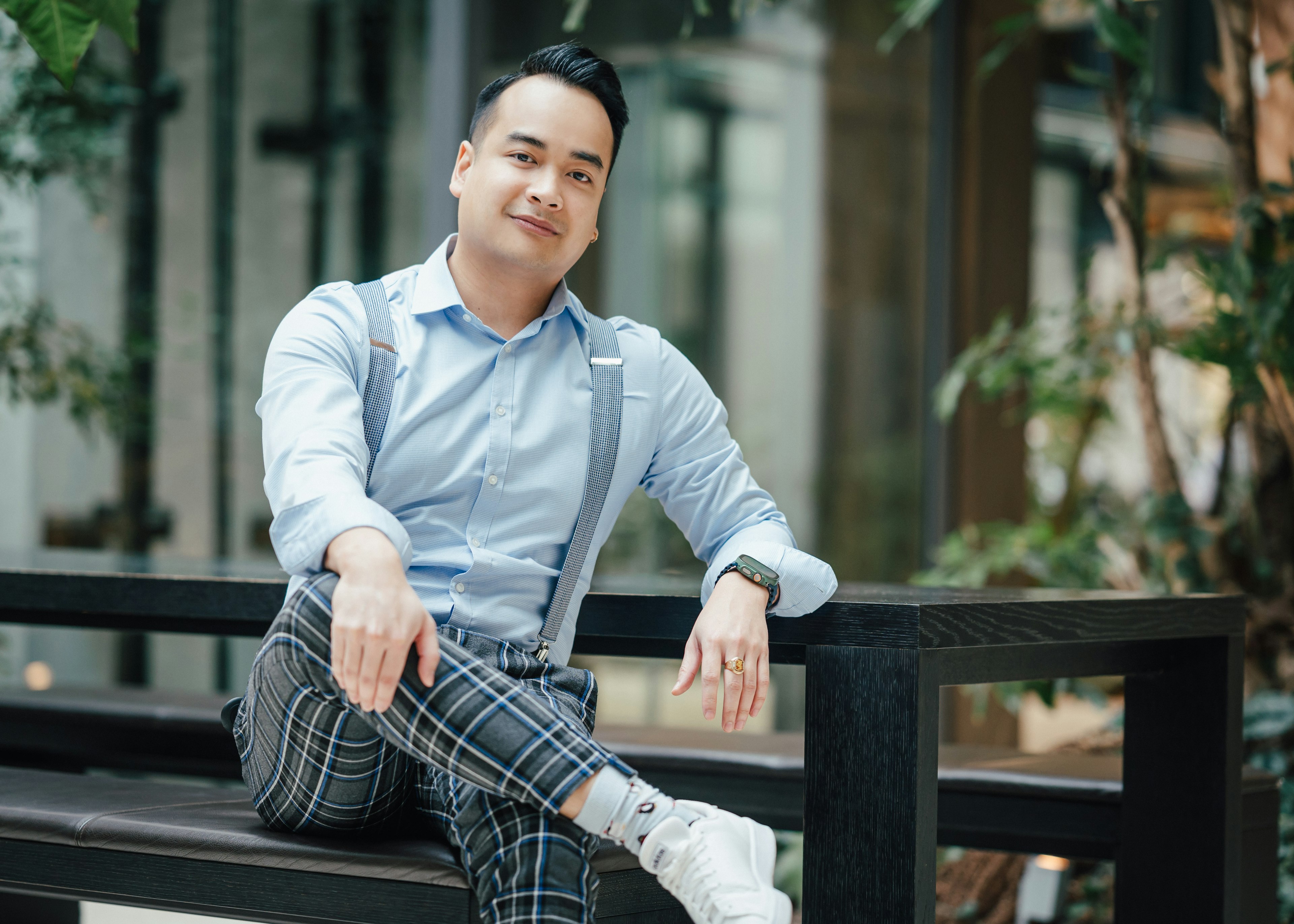 Mann in hellblauem Hemd mit Hosenträgern sitzt entspannt auf einer Bank im begrünten Innenbereich.