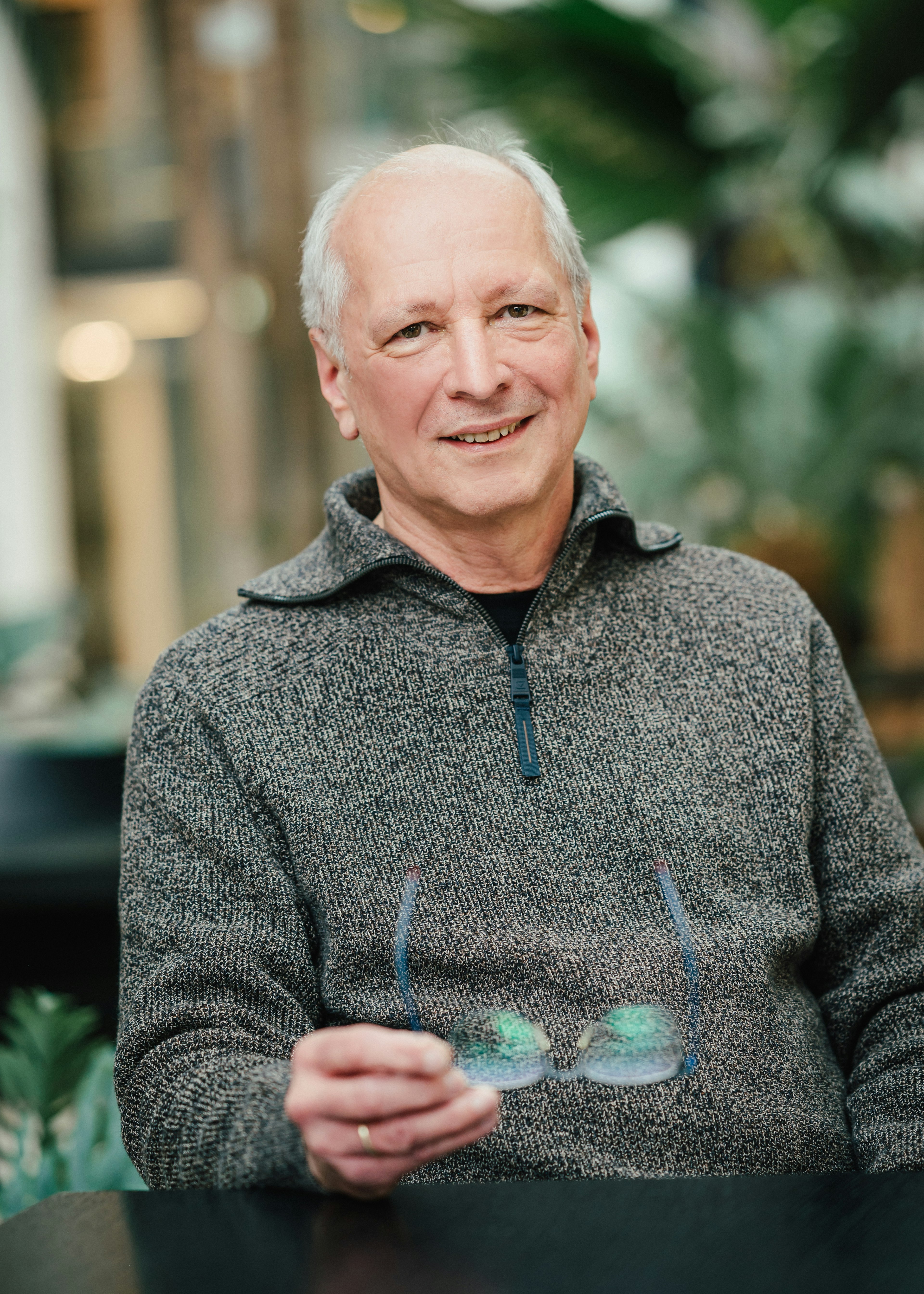 Ein älterer Mann im grauen Pullover sitzt lächelnd am Tisch und hält seine Brille in der Hand.